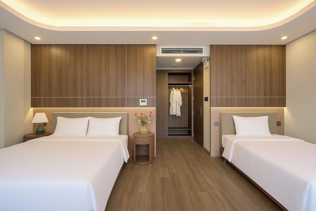A hotel room with two beds, wooden panel walls, a small table with a lamp, and a closet with hanging clothes.