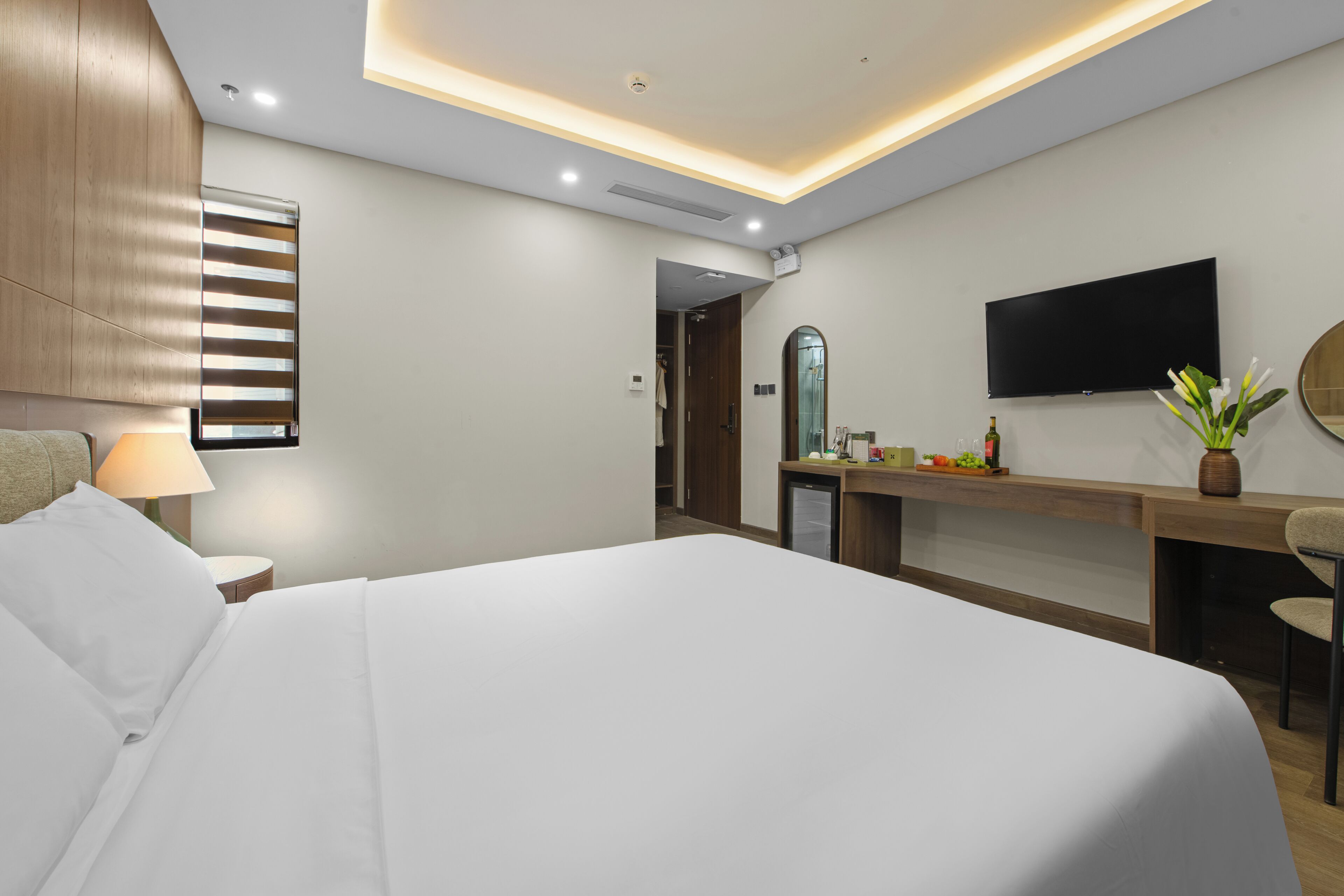 A modern hotel room with a large bed, a desk with a flat-screen TV, a mirror, and a small kitchenette.
