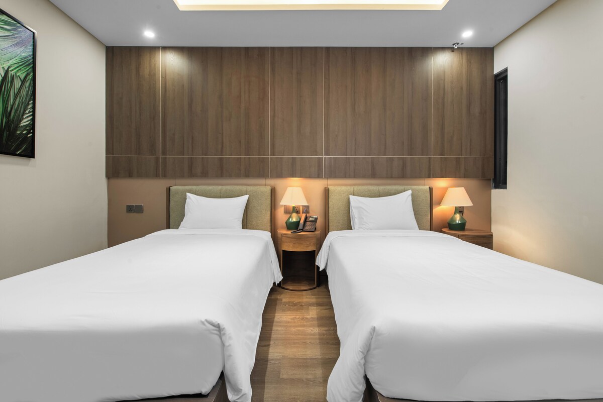 A hotel room with two single beds, wooden headboard, bedside lamps, and a ceiling light.