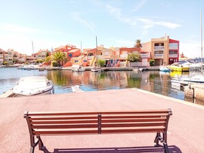 A waterfront marina with boats docked, a bench in the foreground, and colorful buildings in the background. - Odalys Référence Le Lotus Blanc by Interhome (Le Barcarès)