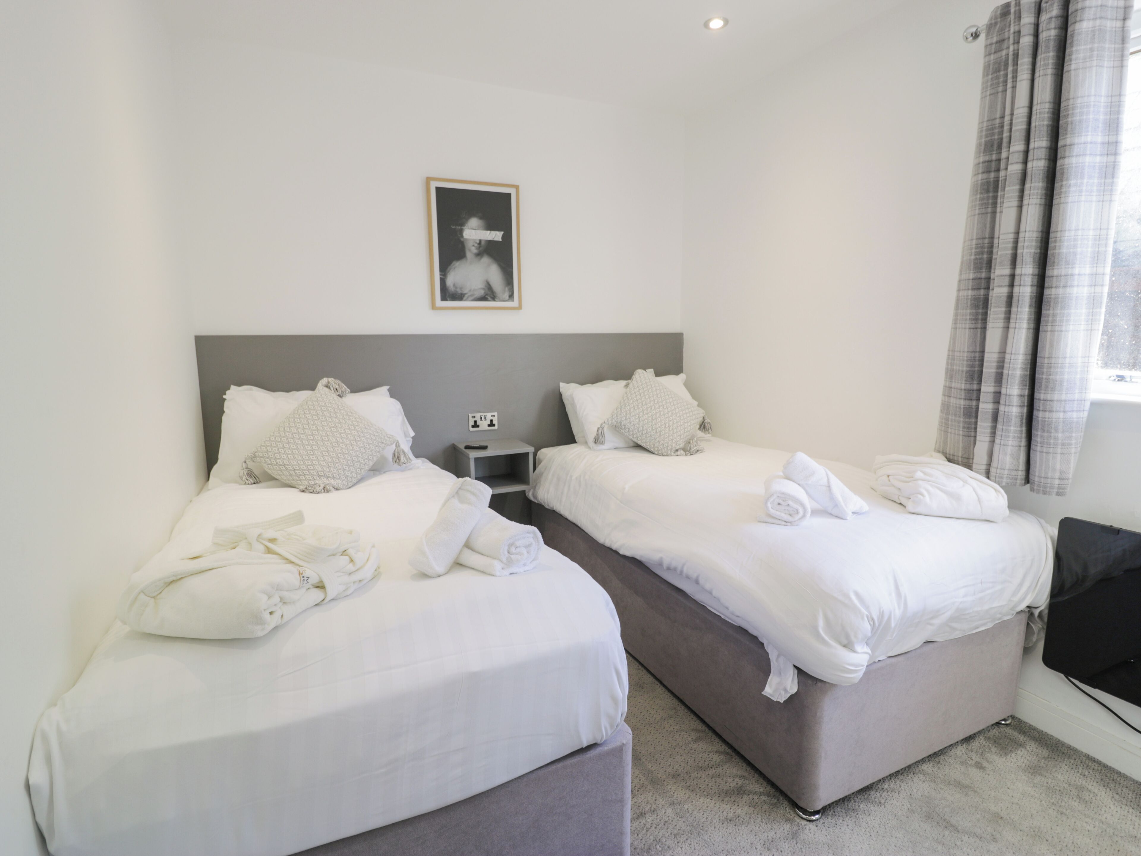 A hotel room with two beds, each with white linens and pillows, a bedside table with a remote control, and a framed picture on the wall.