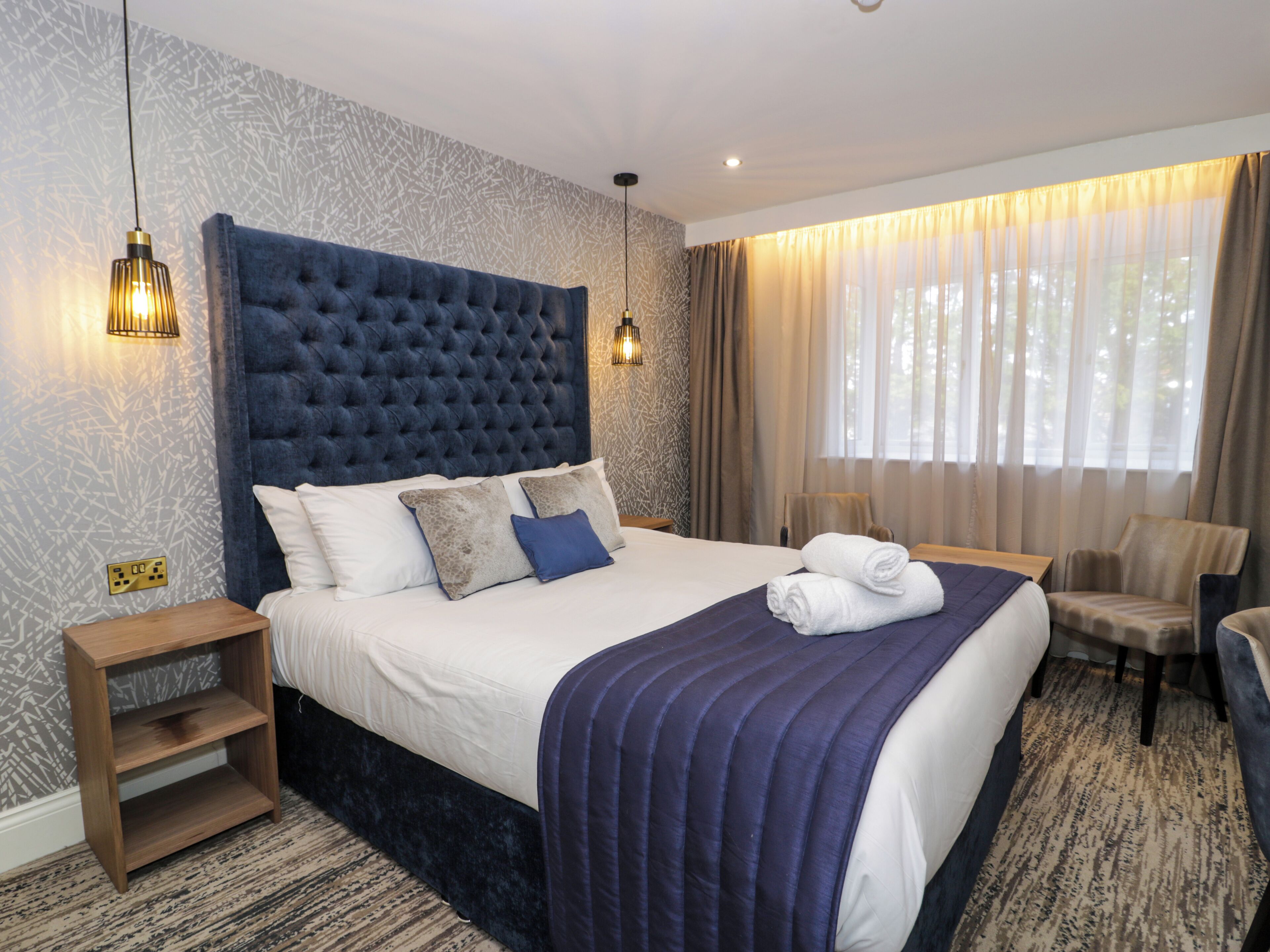 A hotel room with a large bed, a wooden nightstand, a chair, and a window with curtains.