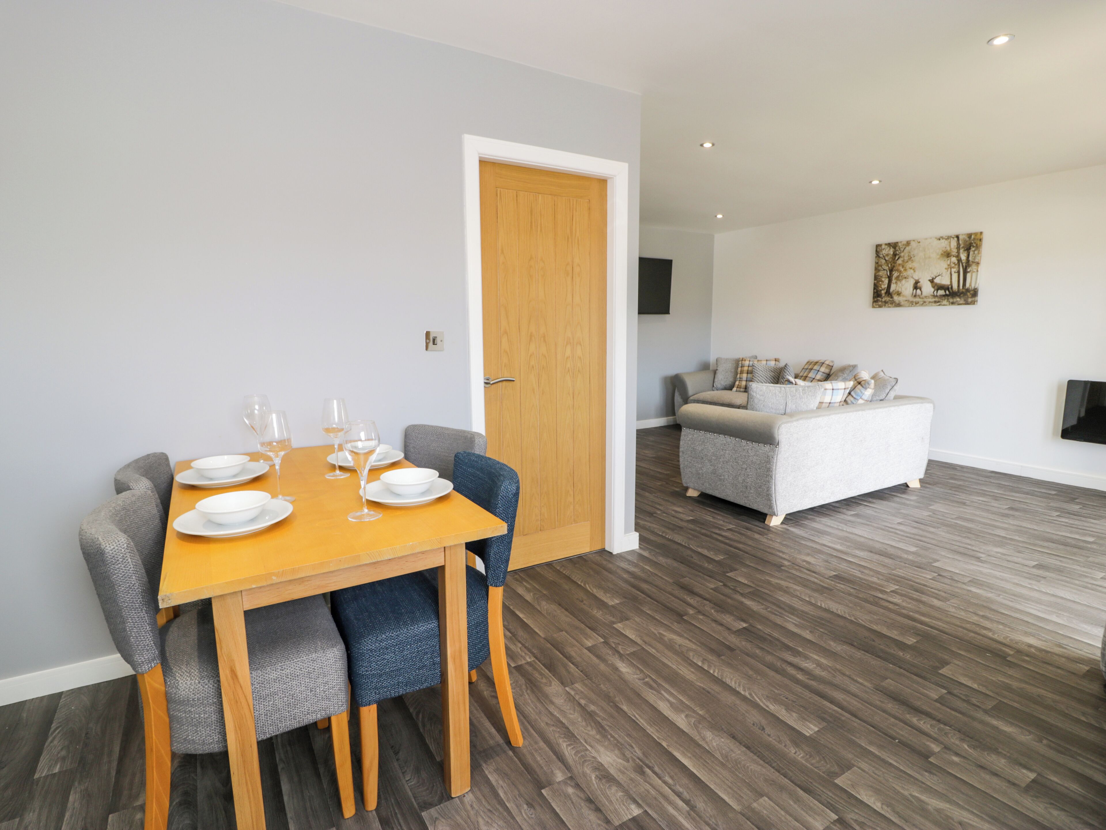A dining area with a wooden table set for four, a grey sofa in the background, and a wooden floor.