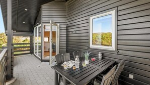 Une terrasse extérieure couverte, dotée d’une table et de chaises, une porte-fenêtre coulissante donnant sur un espace intérieur, et une fenêtre offrant une vue sur la verdure.
