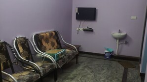 Una habitación con paredes moradas, un ventilador de techo, un televisor de pantalla plana montado en la pared, un lavamanos y dos sillas estampadas.