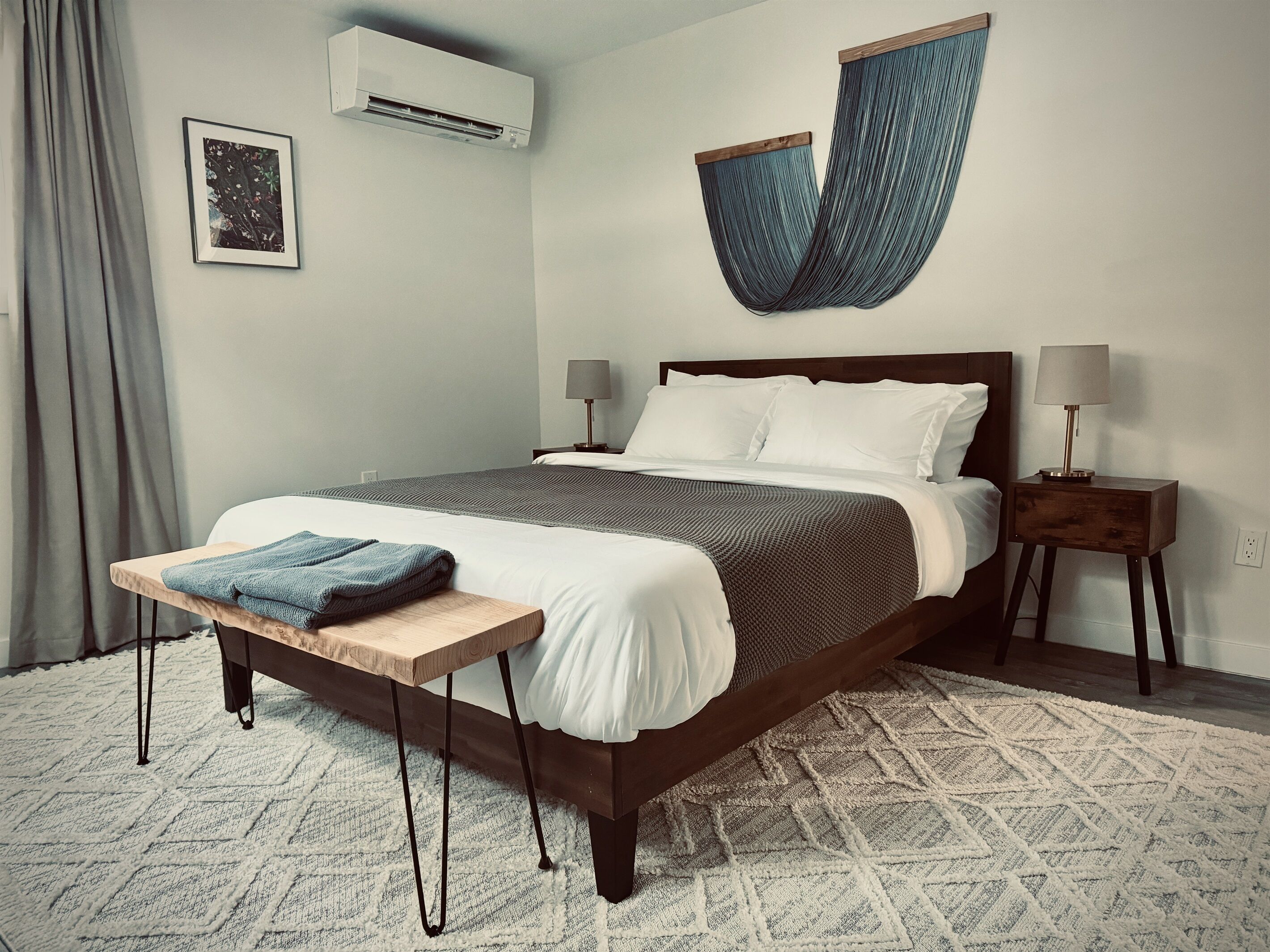 Une chambre à coucher bien rangée, avec un lit, des tables de chevet, un climatiseur fixé au mur et une œuvre textile suspendue.