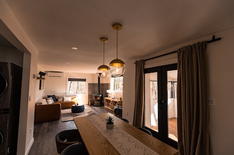 A modern living room with a fireplace, a dining area with a wooden table, and a large window with curtains.