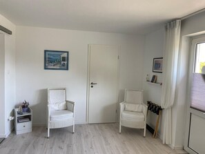 Une piĂšce avec deux chaises blanches, une porte, une petite Ă©tagĂšre avec une plante et une fenĂȘtre avec des rideaux.