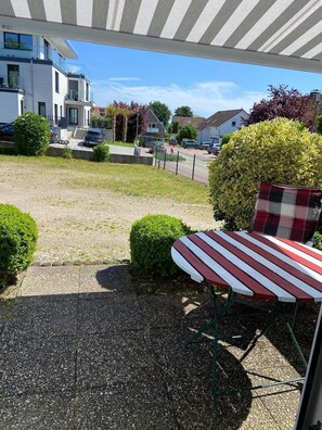 A covered outdoor seating area with a table and chairs, a hedge, and a view of a residential street.