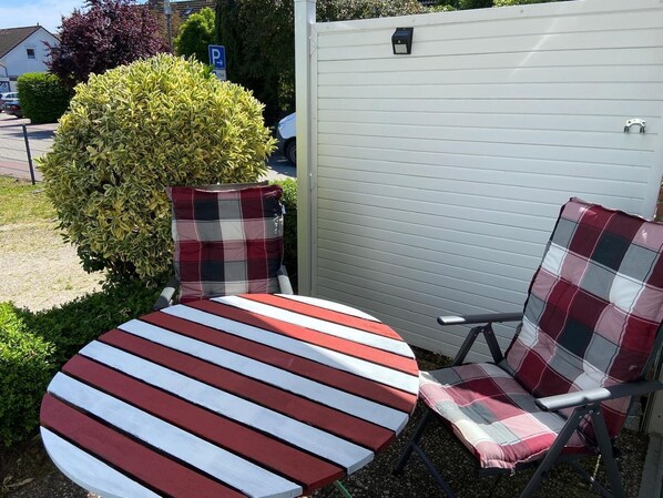 A round table with striped upholstery, two plaid chairs, a small bush, and a white fence with a mailbox.
