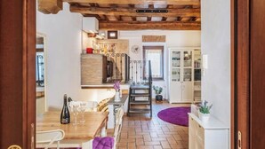 Een rustiek interieur met zichtbare houten balken, een eethoek met tafel en stoelen, een keuken met kasten, en een trap naar een ander niveau.
