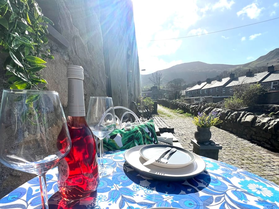 Mesa al aire libre con una botella de vino tinto y una copa, un plato con cubiertos y un mantel estampado.