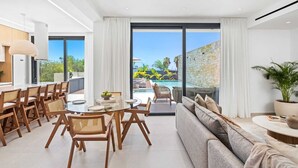 Una sala de estar moderna con una mesa de comedor de vidrio, sillas de madera, un sofá gris y vista a una piscina y follaje.