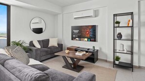 A modern living room with a grey sofa, a coffee table, a TV mounted on the wall, and a bookshelf. - Aether Residence by Nomads (Larnaca)