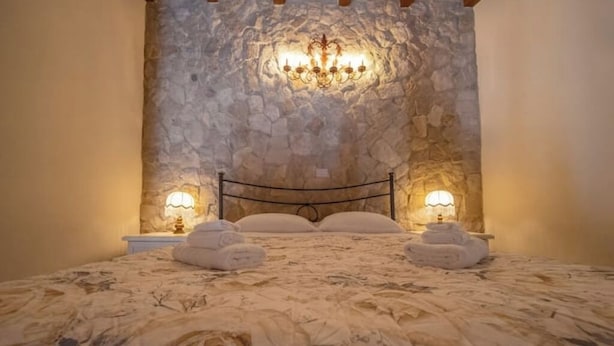 Ein Schlafzimmer mit Steinwand, einem Bett mit blumigem Überwurf, zwei Nachttischlampen und zwei gefalteten Handtüchern auf dem Bett.