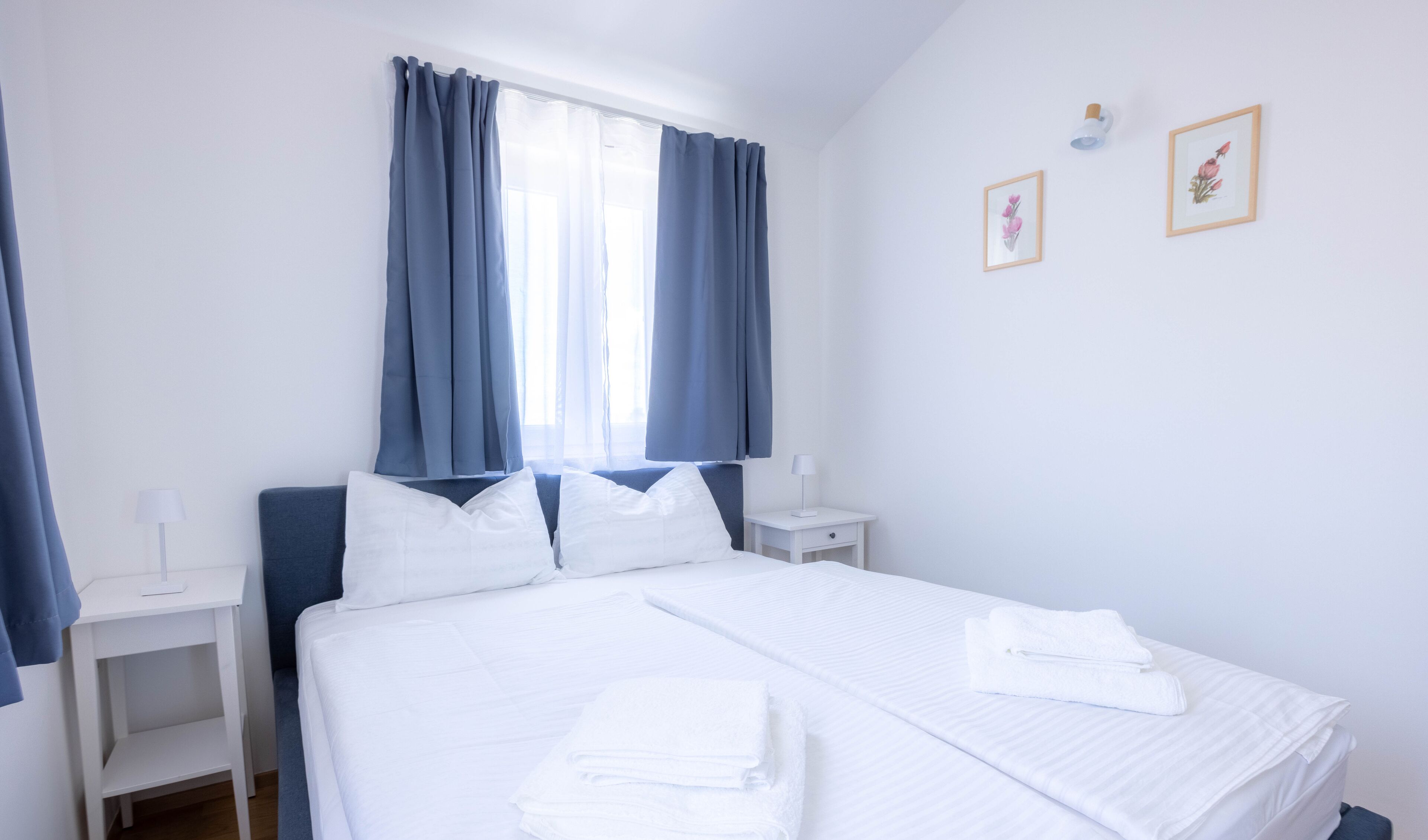 A neatly made bed with white linens and towels, a bedside table with a lamp, and a window with blue curtains.