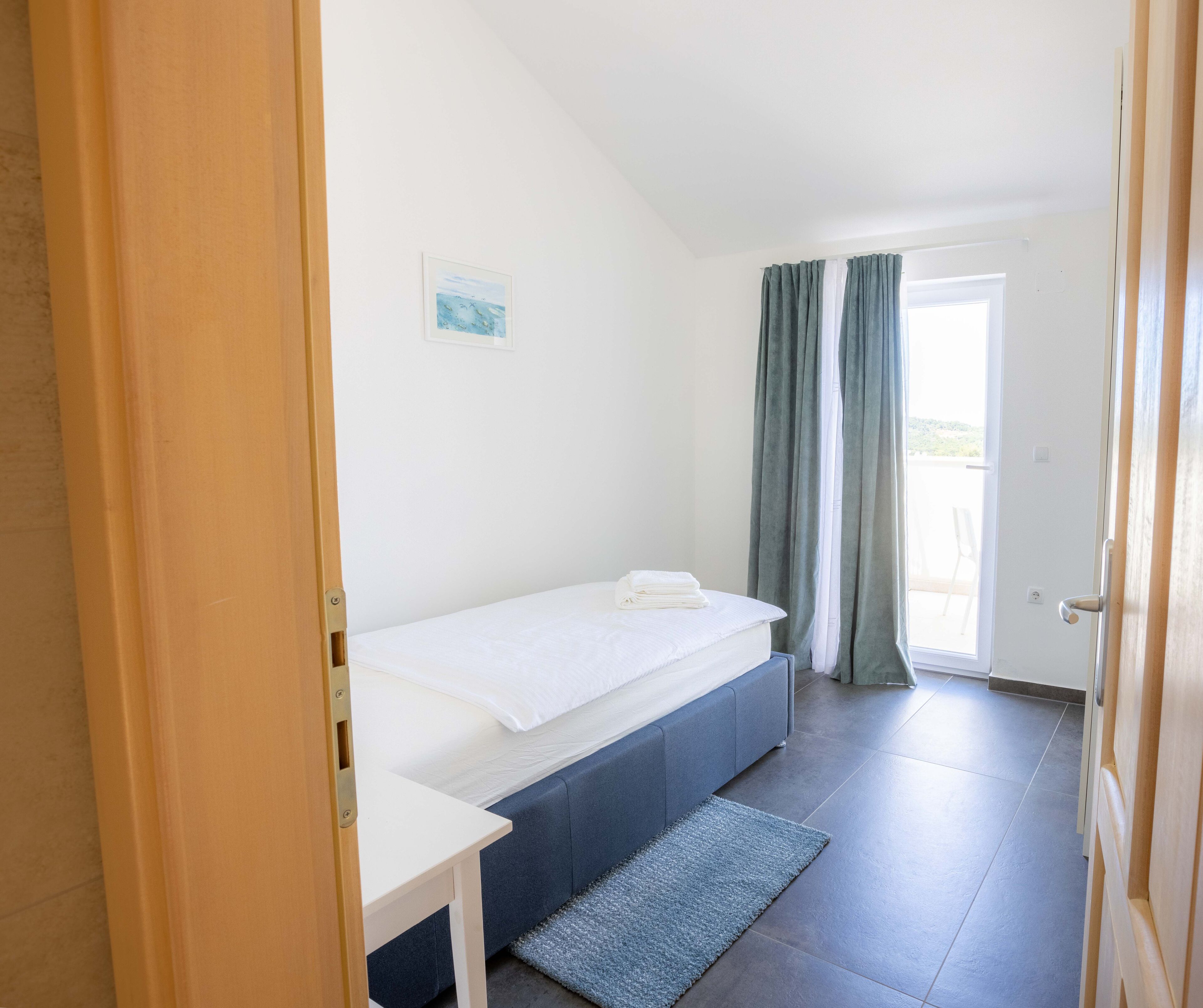 A small, clean room with a single bed, a small table, and a view of the outdoors through a door.