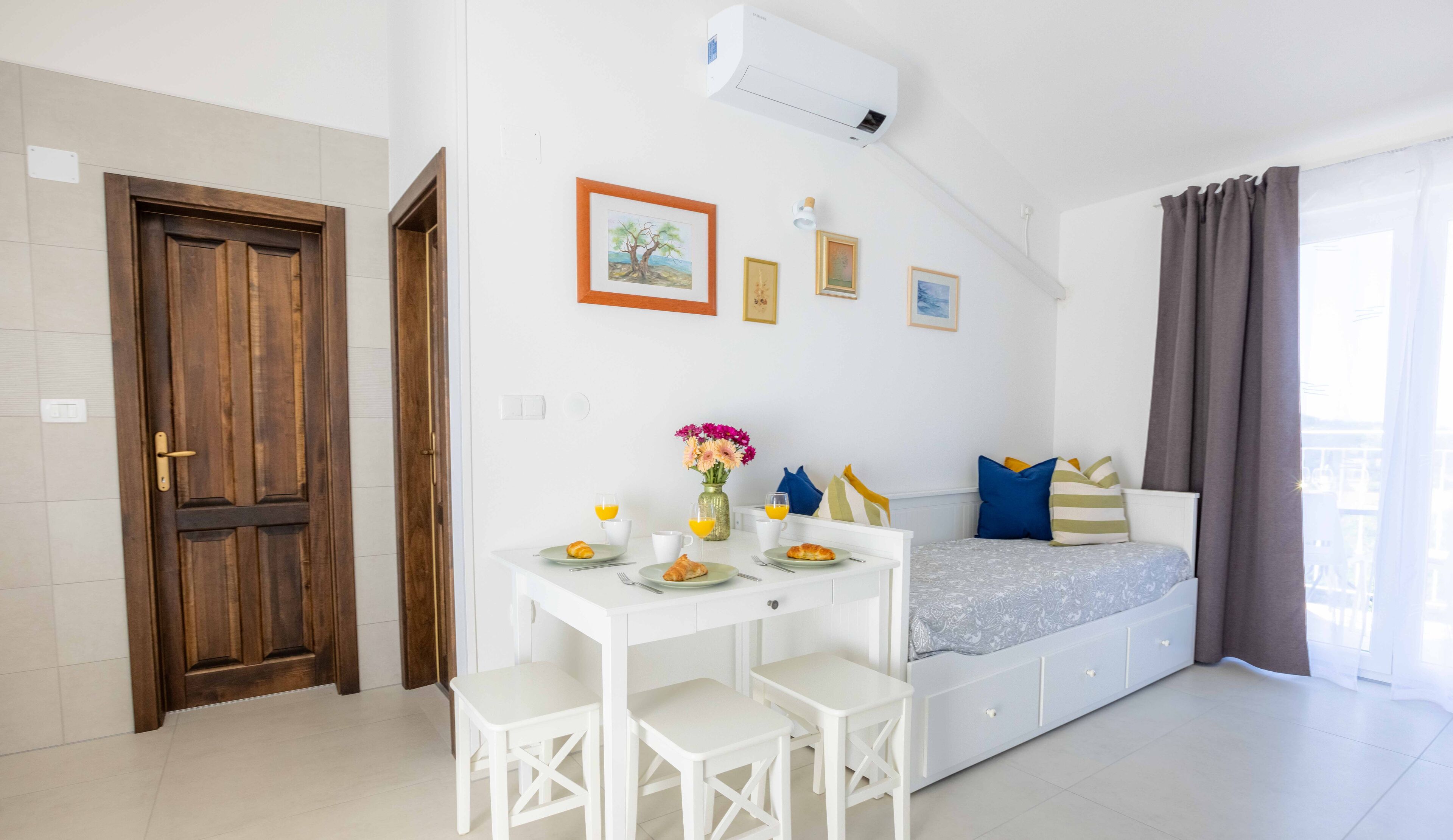 A compact room with a bed, a dining table with chairs, a small white cabinet, and a window with curtains.