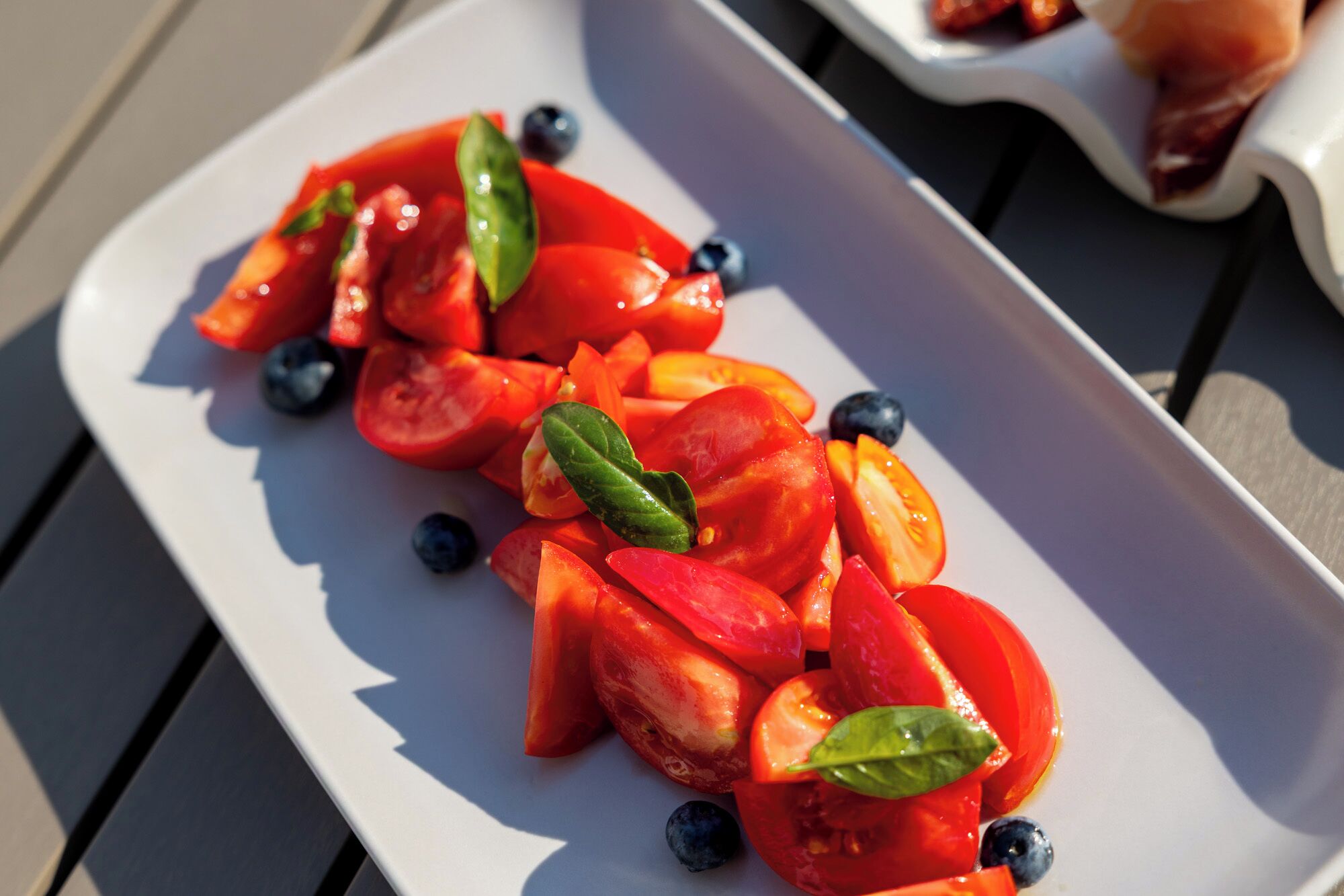 Ein weißer Teller mit Tomatenscheiben, Basilikumblättern und Blaubeeren auf einem Tisch.