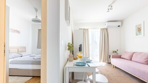Een moderne hotelkamer met een bed, een bank, een eettafel met ontbijtartikelen en een groot raam met gordijnen.