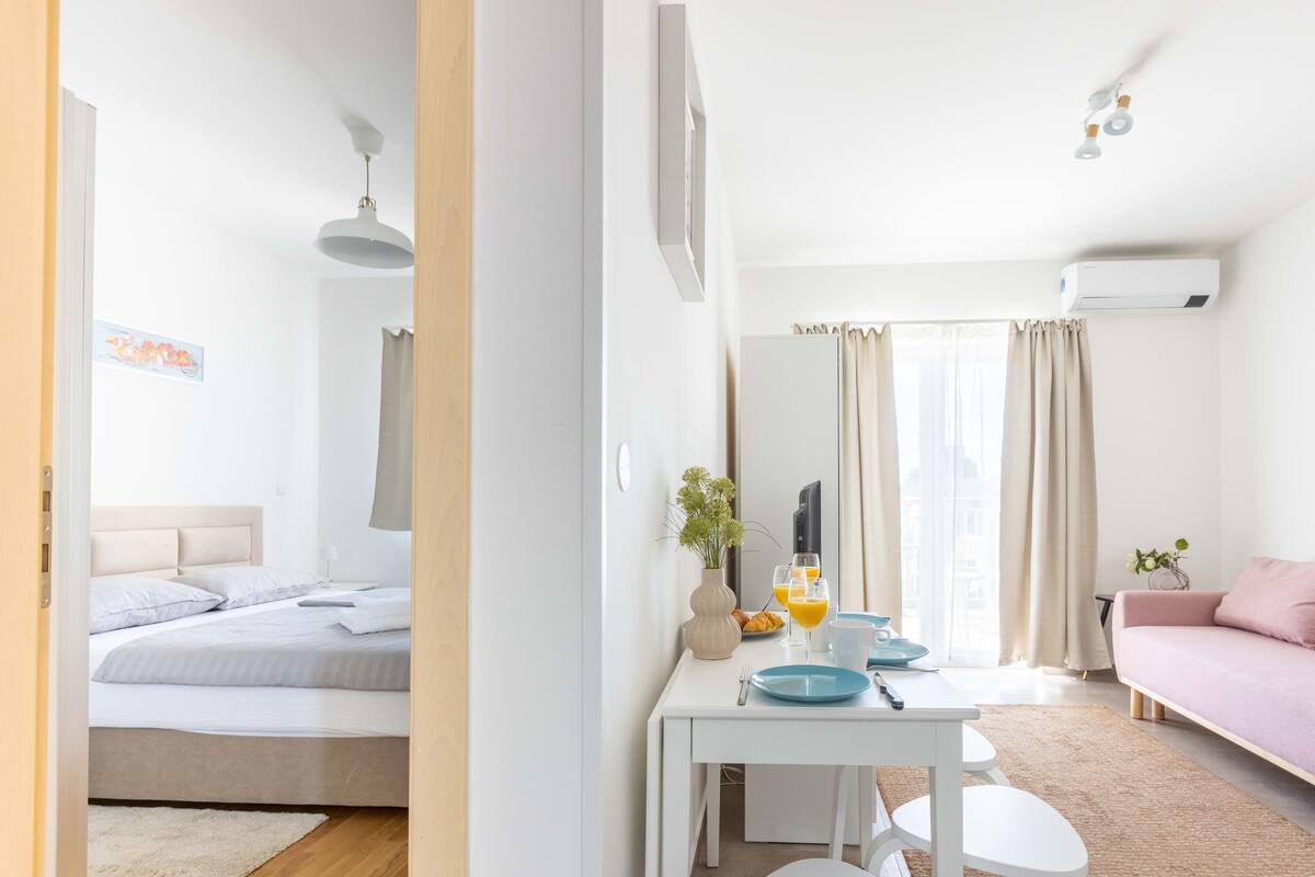 Moderni hotellihuone, jossa on sänky, sohva, aamiaistarvikkeilla katettu ruokapöytä ja suuri ikkuna, jossa on verhot.