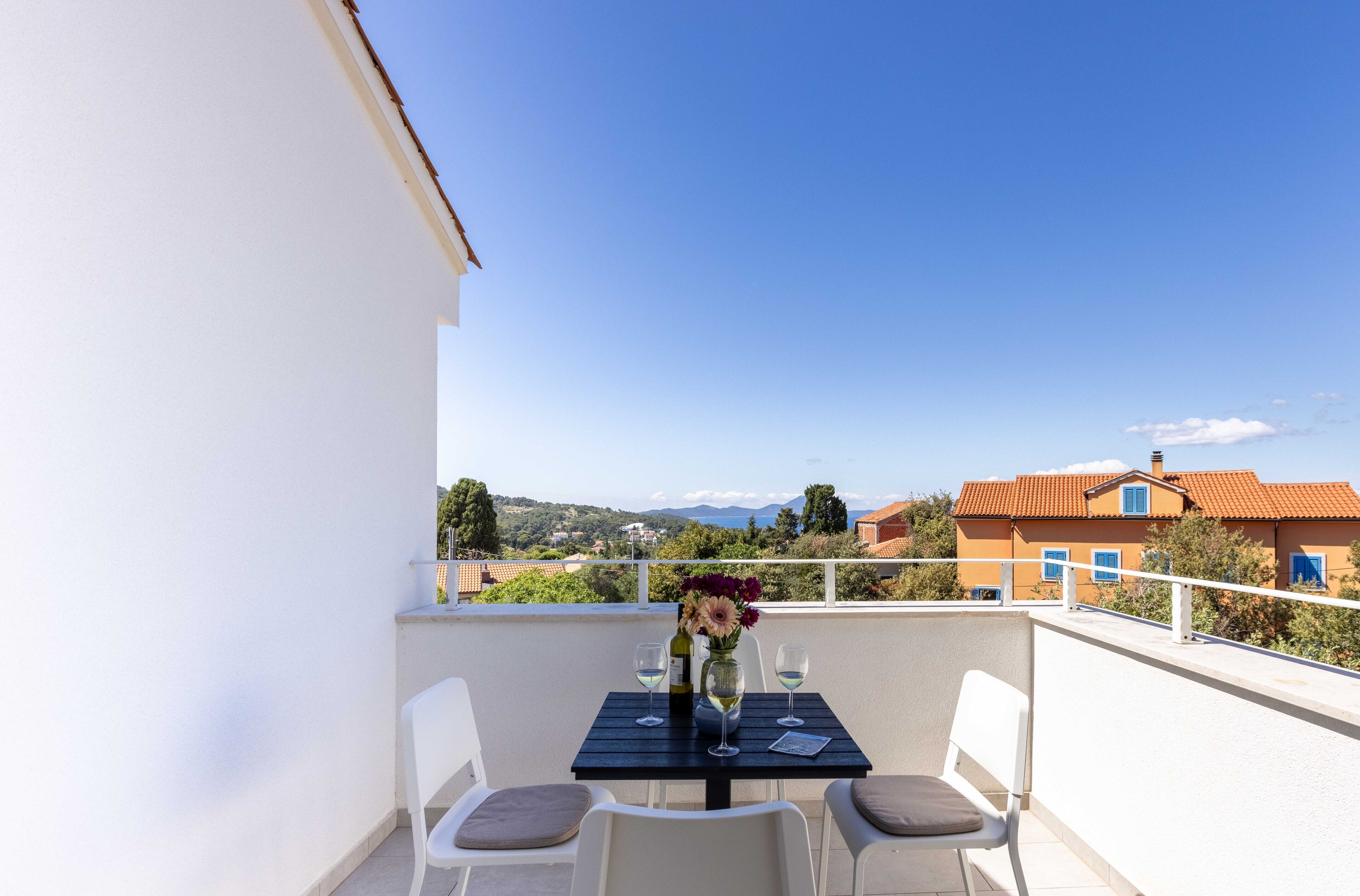 A balcony with a table set for two, a vase with flowers, and a view of a coastal town.