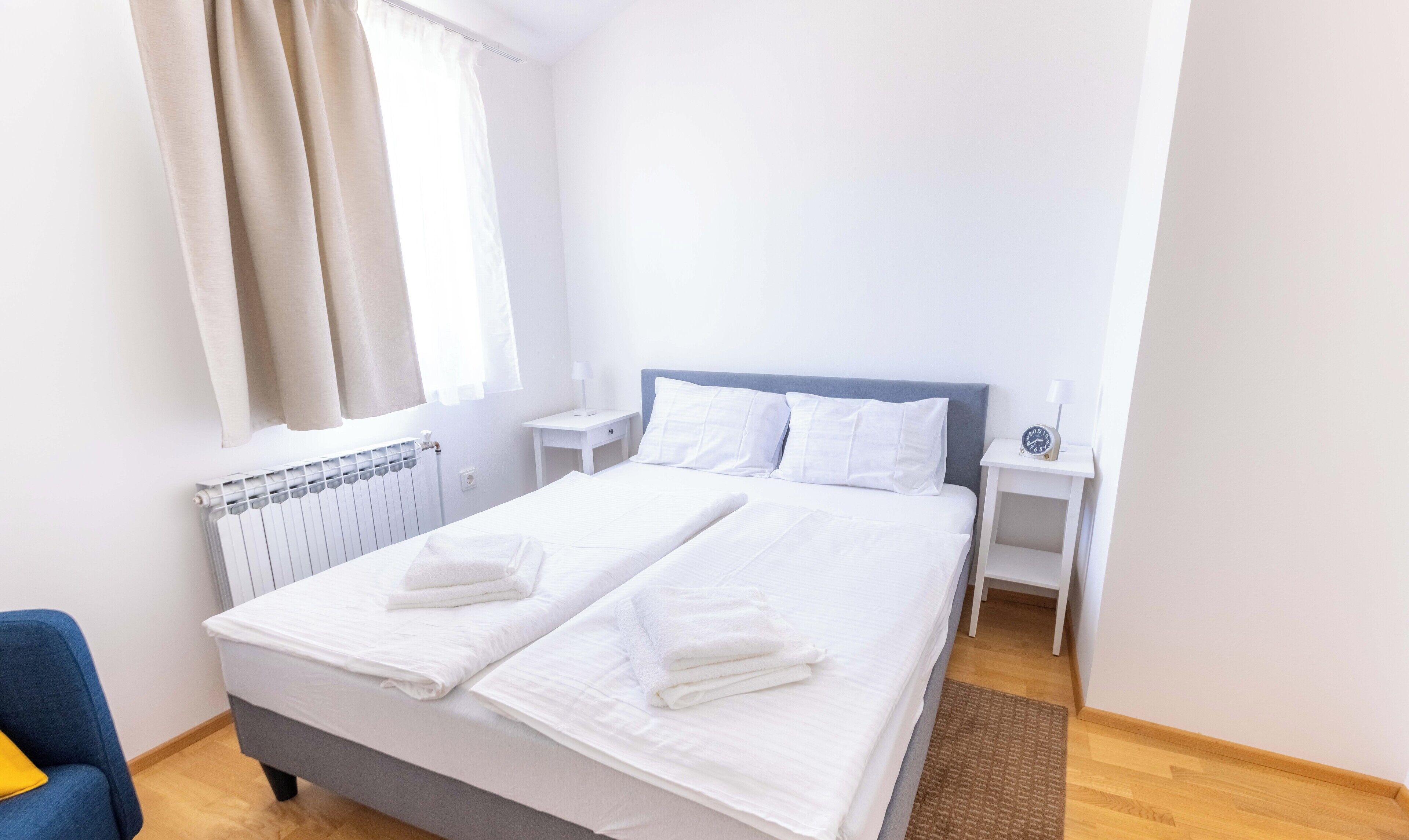 Un lit bien fait, avec du linge de lit blanc et deux serviettes pliées, une table de chevet avec une lampe, une petite horloge et une fenêtre avec des rideaux.