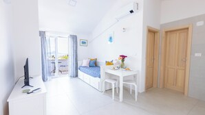 Een lichte, moderne kamer met een bed, een bureau en een stoel. Er is een raam met gordijnen, een wandairconditioner en een deur.