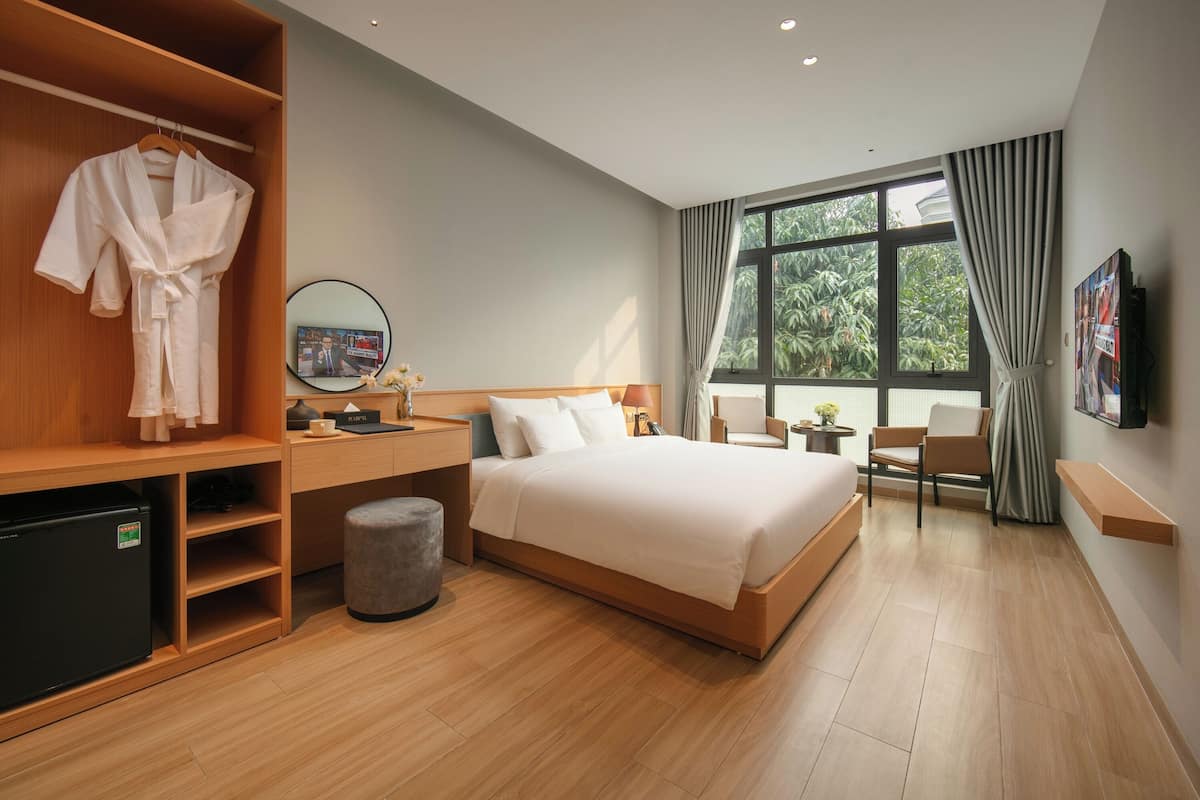 A modern hotel room with a large bed, a wooden wardrobe, a desk with a computer, and a small seating area.
