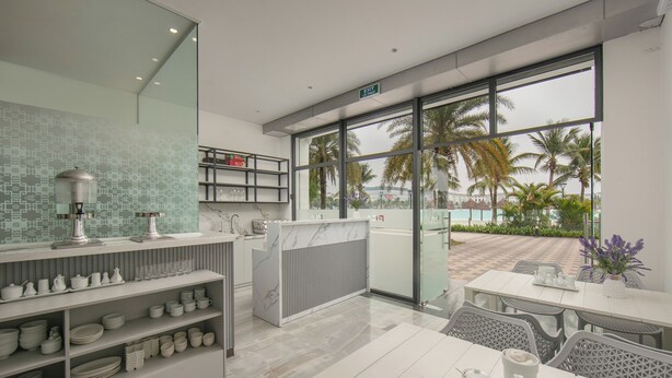 A modern hotel room with a large window, a balcony with a view of palm trees, and a neatly organized kitchen area. - PCA Hotel 1- Vinhomes Ocean Park (Hanoi)