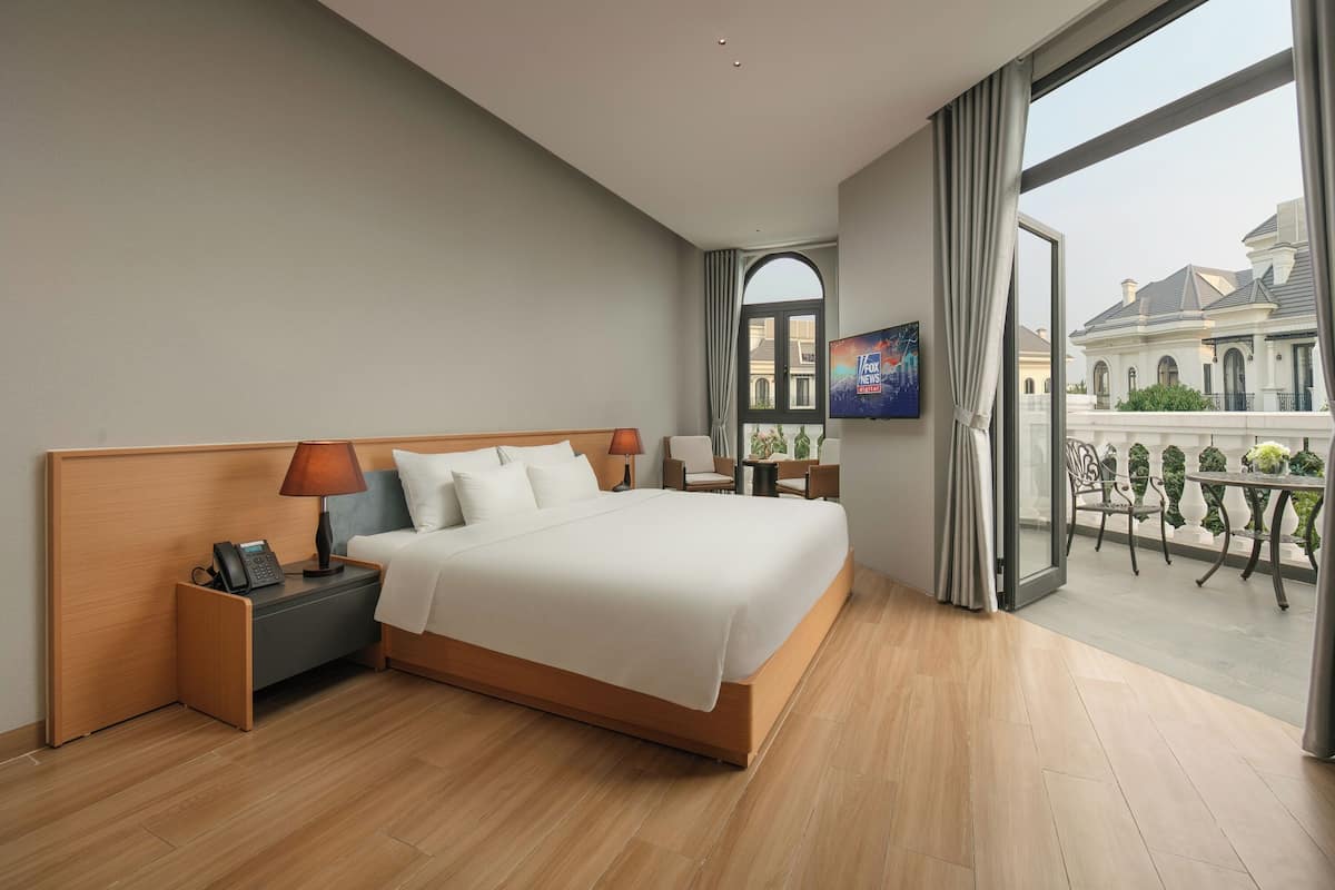 A modern hotel room with a large bed, a nightstand with a phone, a TV on the wall, and a balcony with a view of a building.