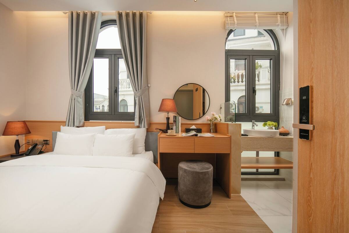 A hotel room with a large bed, a desk with a mirror, a vanity with a sink, and a small stool.