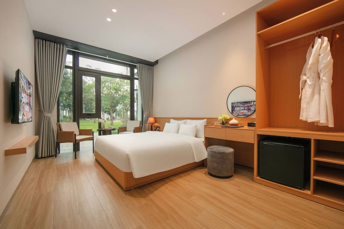 A modern hotel room with a large bed, a wooden wardrobe, a small desk, a chair, a TV, and a window with curtains.