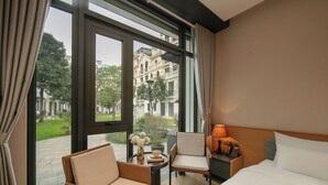 A hotel room with a bed, a chair, a small table with a lamp, and a view of the outside. - PCA Hotel 1- Vinhomes Ocean Park (Hanoi)