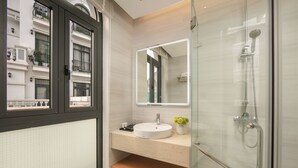 A modern bathroom with a glass-enclosed shower, a large mirror, and a window overlooking a building. - PCA Hotel 1- Vinhomes Ocean Park (Hanoi)