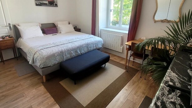 A bedroom with a bed, bedside table, nightstand, window with curtains, radiator, and a mirror on the wall.