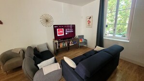 A living room with a dark blue sofa, a grey armchair, and a bookshelf with books and decorative items. A television is mounted on the wall, and there is a window with curtains.