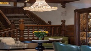 A grand staircase with wooden railings and a chandelier, a green sofa, a patterned armchair, and a table with a vase of flowers.