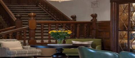 A grand staircase with wooden railings and a chandelier, a green sofa, a patterned armchair, and a table with a vase of flowers.