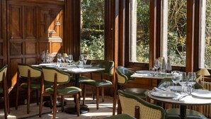 A dining area with wooden paneled walls, large windows with a view of greenery, and green upholstered chairs.