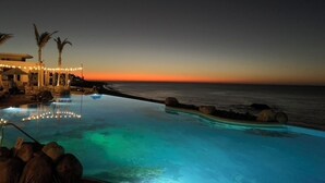 Une piscine à débordement au coucher du soleil, avec vue sur l’océan, des palmiers et un bâtiment illuminé de guirlandes lumineuses.