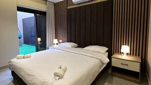 A modern hotel room with a large bed, two bedside tables with lamps, a ceiling-mounted air conditioner, and a view of a green outdoor area. - 004 AL-NAJEH JRE Apartments For Rent ave (Amman)