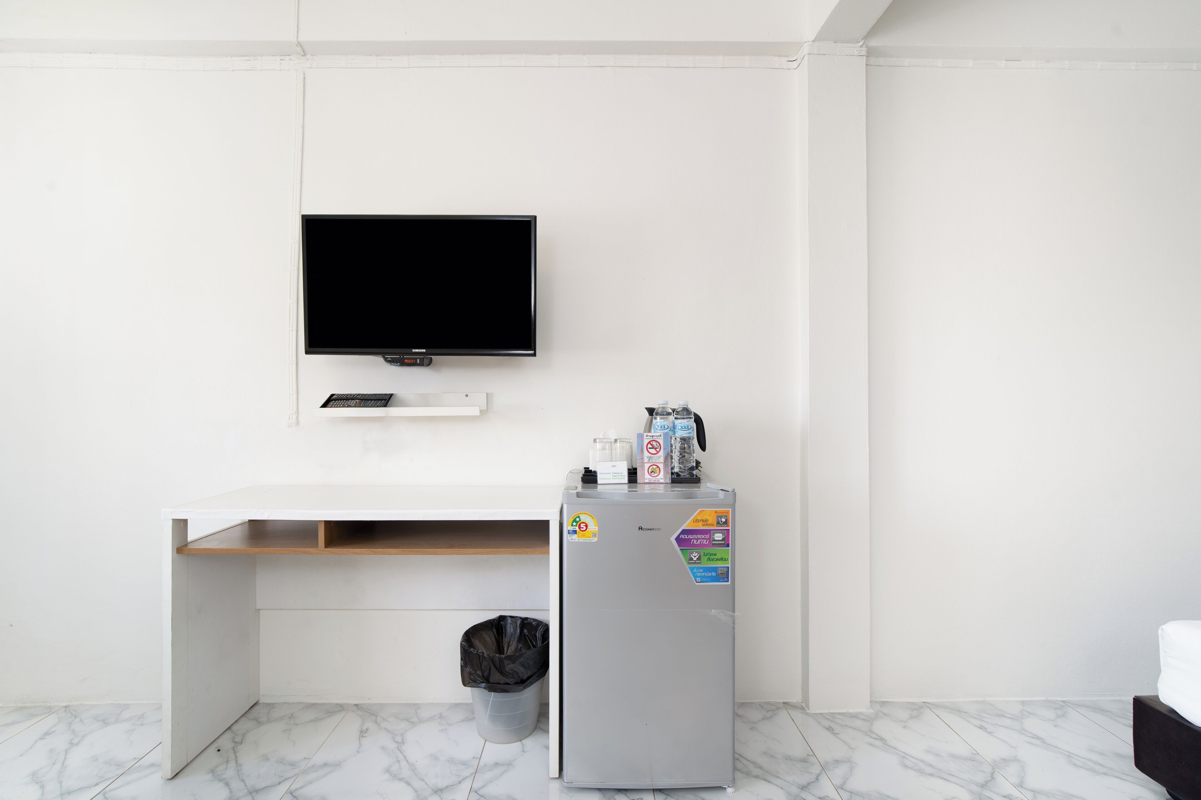A modern room with a flat-screen TV mounted on the wall, a small refrigerator with recycling stickers, a trash bin, and a wooden shelf under a white desk.