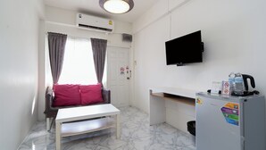 A compact room with a white table, a sofa with a red cushion, a television mounted on the wall, a refrigerator with various stickers, and a window with curtains.