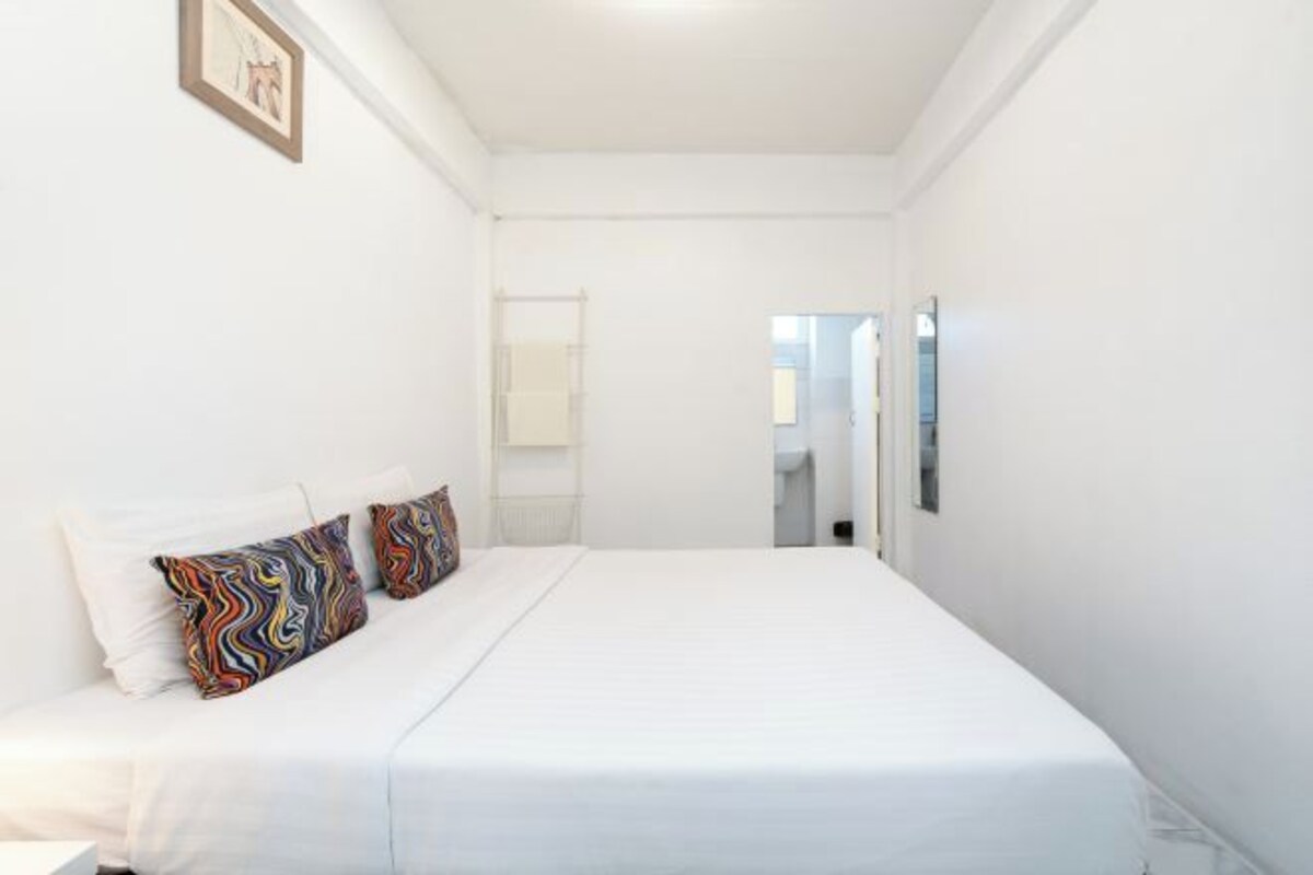 A white room with a bed, a framed picture on the wall, and a mirror on the wall.