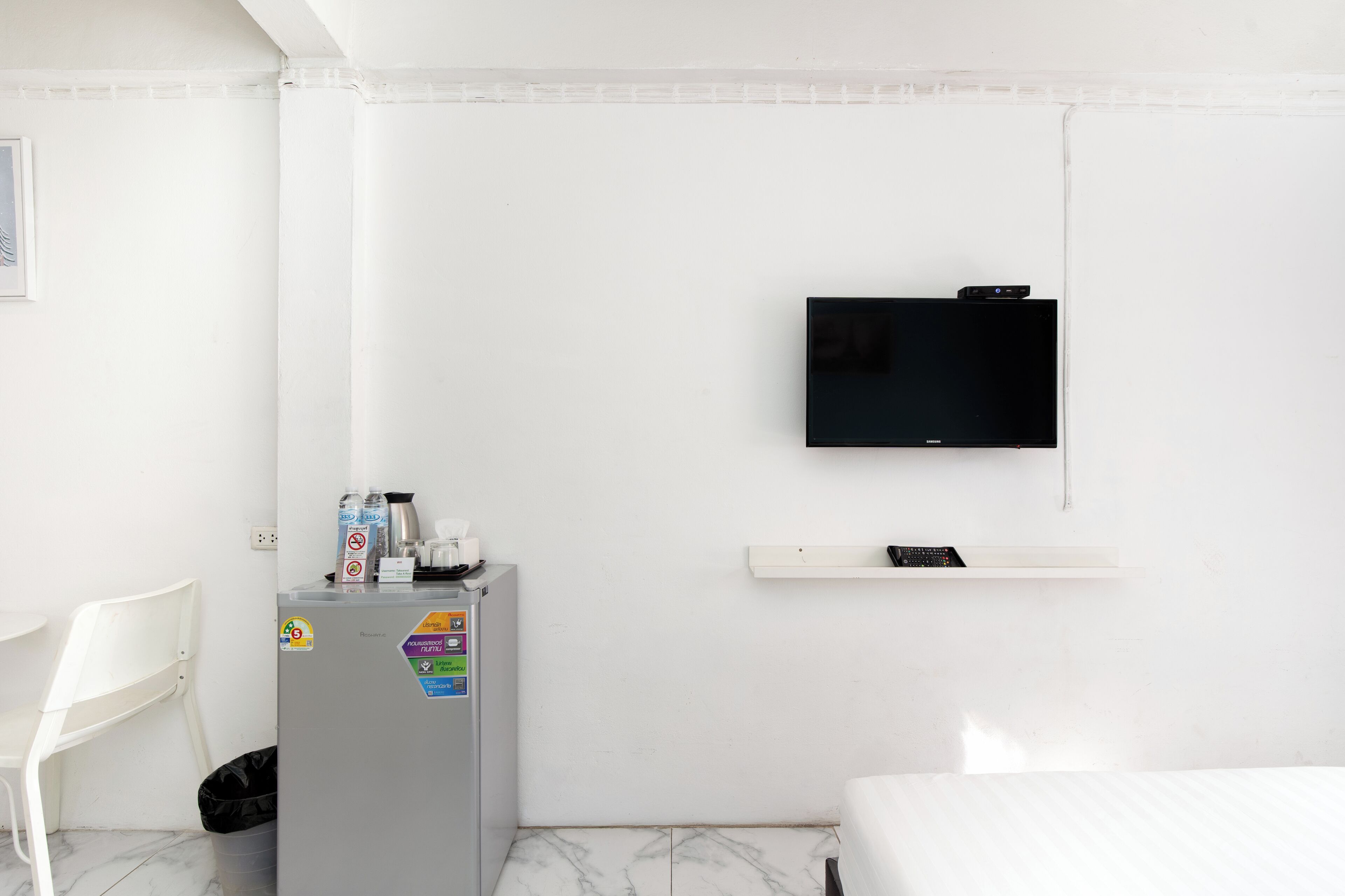 Habitación de hotel con cama, televisión montada en la pared, un refrigerador pequeño, una silla y un bote de basura.