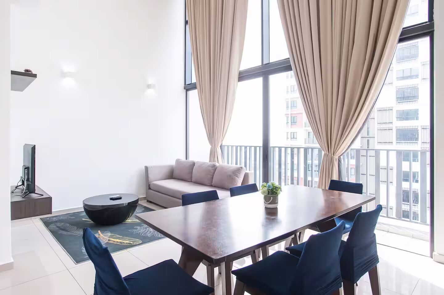 Una sala de estar moderna con un sofá, una mesa de comedor con sillas, un televisor sobre un mueble bajo y ventanas grandes con cortinas.
