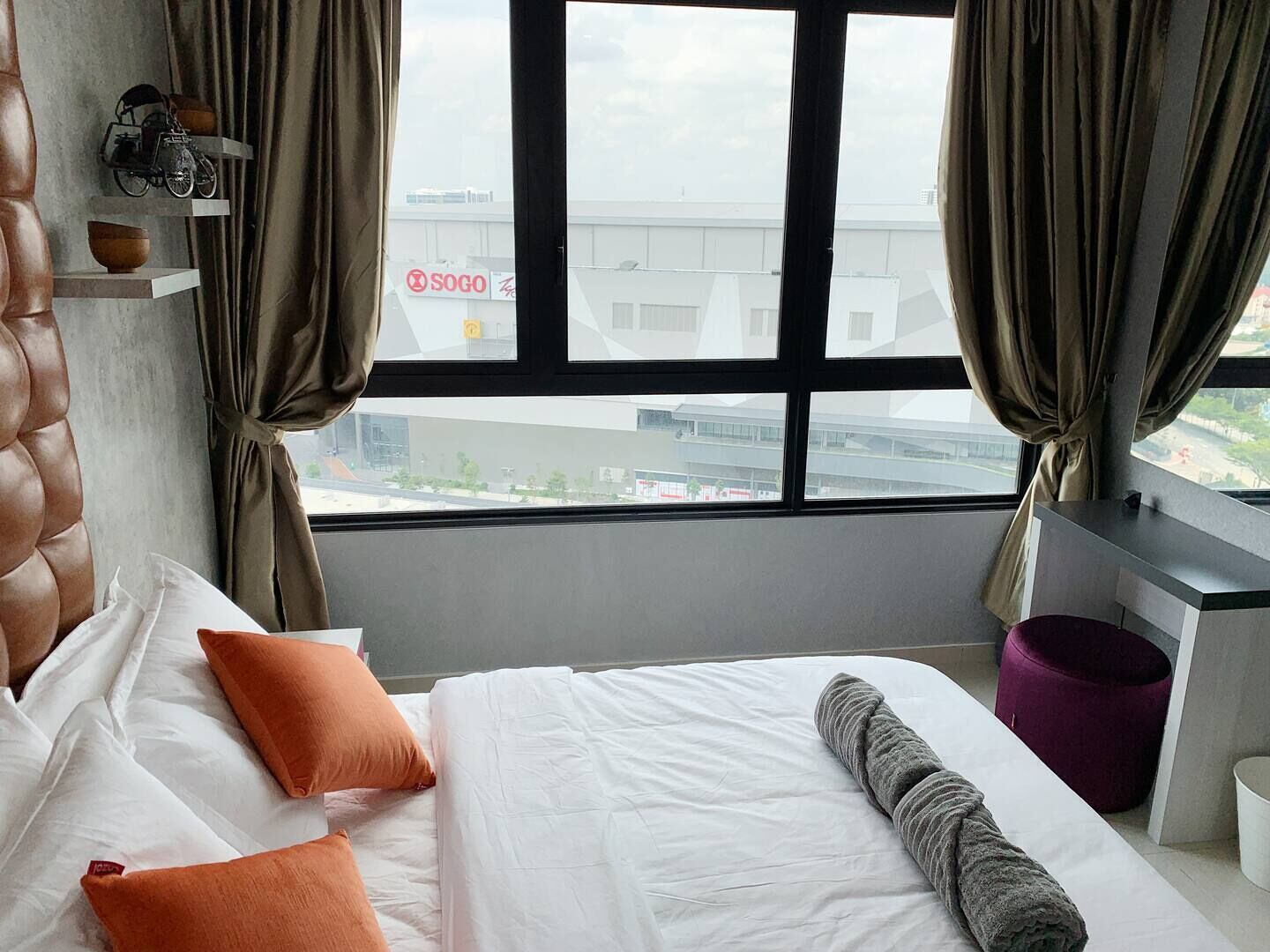 A hotel room with a large window, a bed with white bedding and orange pillows, a grey towel, and a purple ottoman.