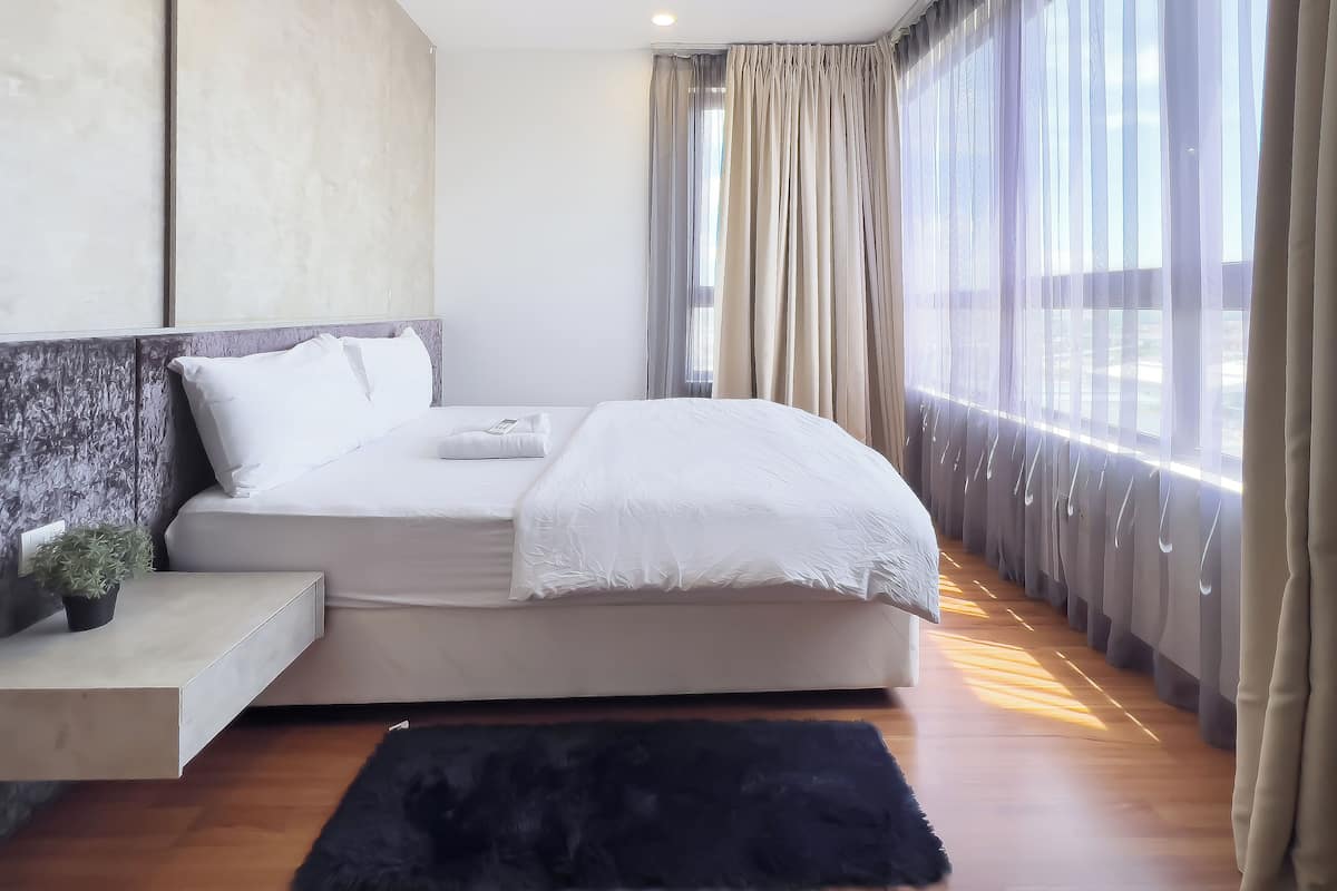 Une chambre à coucher moderne avec un grand lit, une table de chevet agrémentée d’une plante et une vue sur l’extérieur à travers des rideaux légers.