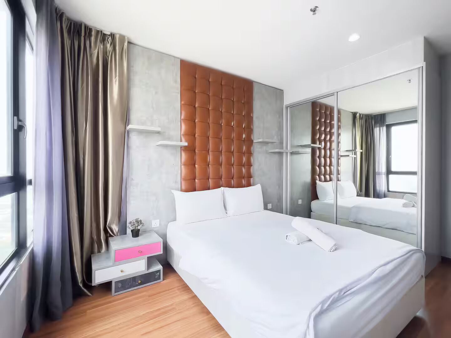 Een moderne hotelkamer met een groot bed, een nachtkastje met een roze lade, een spiegel en uitzicht op de stad.